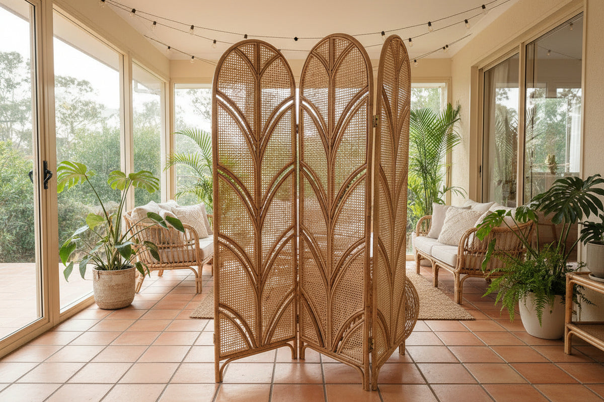 A three-panel folding room divider made of natural woven rattan or wicker, standing in a bright, modern bedroom/living space between a sofa and a bed.